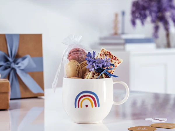 Une tasse « With Love » de Villeroy & Boch au design arc-en-ciel remplie de macarons Une tasse « With Love » de Villeroy & Boch au design arc-en-ciel, remplie de macarons, d’une tablette de chocolat et avec fleurs violettes sur une surface blanche avec des boîtes cadeaux et des livres en arrière-plan.