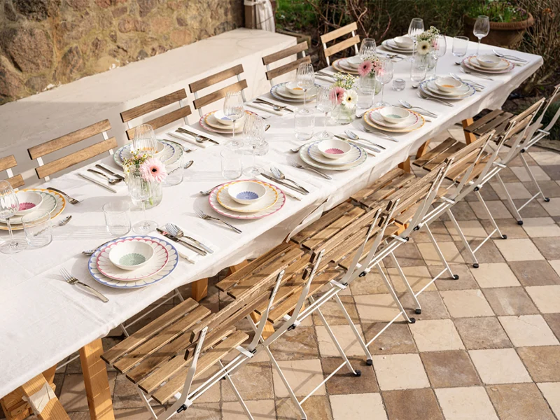 Une longue table à manger en plein air est dressée avec des assiettes, des verres, des couverts et des fleurs sur une terrasse à carreaux à côté d'un mur en pierre