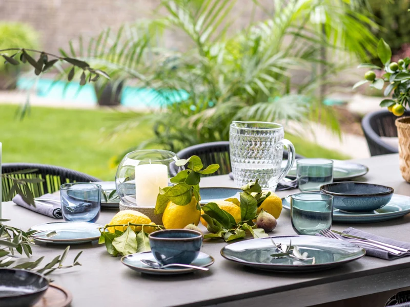 Une table de repas en plein air dressée avec de la vaisselle Lave, des verres, des bougies et des agrumes frais en guise de centre de table, avec vue sur une piscine.