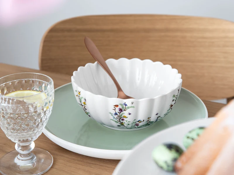 Un bol décoratif avec un motif floral de la collection Spring Fantasy de Villeroy & Boch. Un bol décoratif avec un motif floral de la collection Spring Fantasy de Villeroy & Boch et une cuillère sur une assiette verte. Un verre d'eau avec du citron et une assiette de macarons variés sont disposés à côté sur la table.