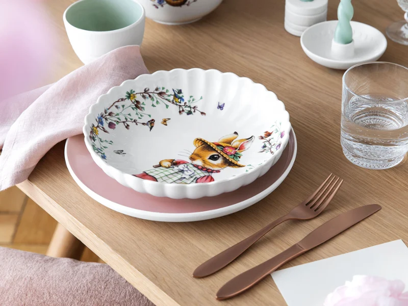 Le bol en céramique "Spring Fantasy" avec un motif de lapin de Villeroy & Boch. Le bol en céramique "Spring Fantasy" avec un motif de lapin de Villeroy & Boch est posé sur des assiettes roses et blanches, accompagné de couverts en or rose et d'un verre d'eau sur une table en bois.
