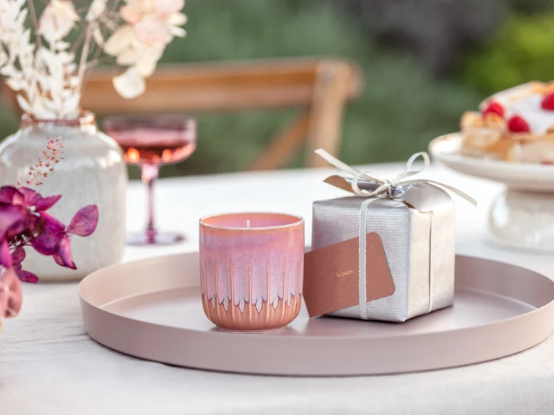 Une bougie parfumée rose Perlemor dans un emballage cadeau Une bougie parfumée de la collection de like. by Villeroy & Boch sous forme de cadeau sur un plateau rond avec une boîte cadeau argentée en arrière-plan et des orchidées roses au premier plan.