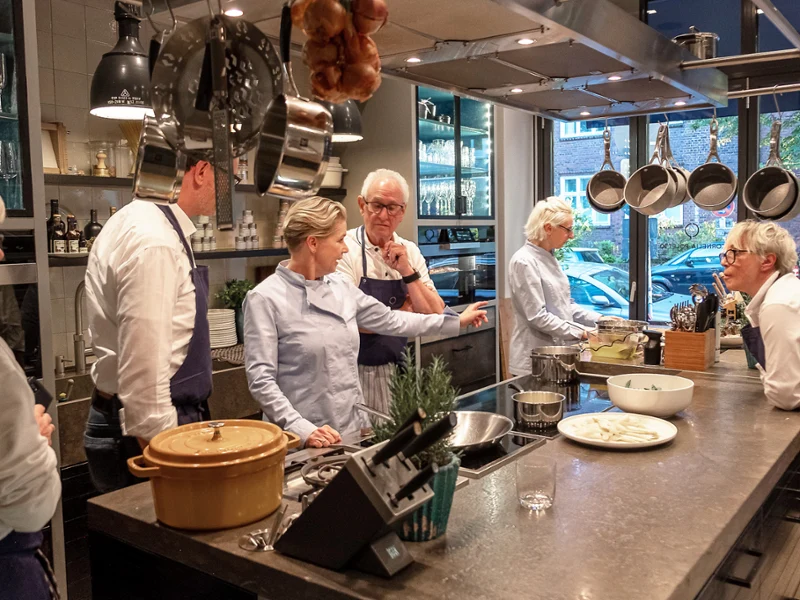 Cinq personnes discutent de cuisine dans une cuisine moderne, debout autour d’un comptoir avec des poêles, des assiettes et des ustensiles.