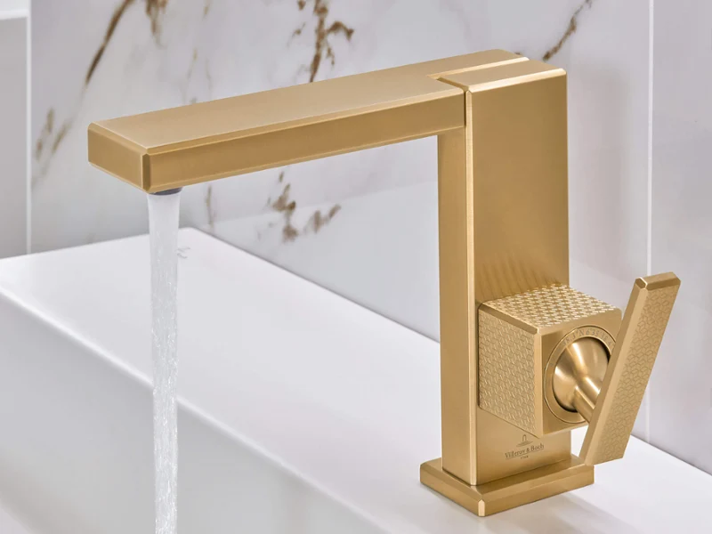 Mitigeur de lavabo Mettlach doré avec eau courante Un robinet doré moderne dans la salle de bains avec de l’eau qui coule devant un dosseret blanc et marbré.