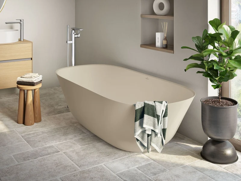 Une salle de bains moderne avec une baignoire en îlot Theano Villeroy & Boch de couleur beige, un tabouret en bois avec des serviettes, une plante en pot et une fenêtre qui laisse passer la lumière du jour. Un sol carrelé et des éléments de design minimalistes.