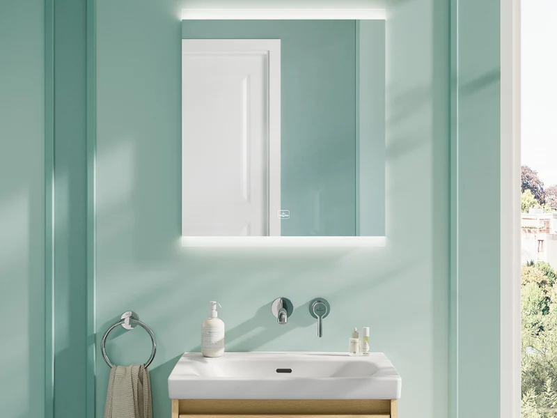 Salle de bain moderne et fraîche avec lavabo Skyla et miroir More To See Square Une salle de bain lumineuse et contemporaine dotée d'un lavabo Villeroy & Boch Skyla avec un meuble sous-vasque en bois clair, associé à un miroir éclairé carré More To See Square. L'espace comprend des murs lambrissés vert menthe doux, un sol en terrazzo, des fixations murales et des accessoires minimalistes.
