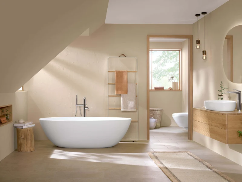 Une baignoire en îlot Antao avec vasque à poser et des robinetteries dans une salle de bains moderne Un intérieur de salle de bains moderne avec une baignoire en îlot, un meuble en bois avec une vasque à poser, des toilettes, des miroirs ronds et des murs beige, avec de la lumière naturelle qui entre par une porte ouverte.