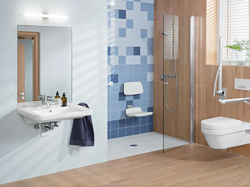 Une salle de bains accessible ViCare avec un siège de douche Une salle de bains accessible avec un sol en bois et un revêtement en bois au niveau du mur. Des poignées rabattables sont montées à côté des toilettes et un siège de douche avec dossier est installé dans la douche.