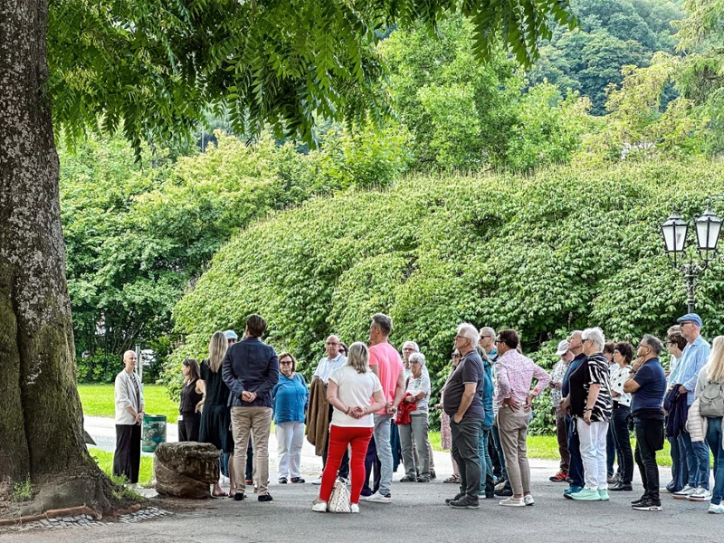 Un groupe de personnes se tient en plein air près d’un grand arbre et d’un lampadaire au milieu du parc Villeroy & Boch, entouré de verdure.