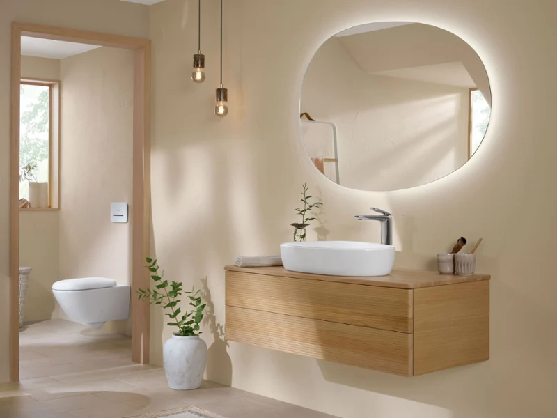 Une salle de bains moderne et lumineuse avec un miroir rond rétroéclairé au-dessus d’un plan de toilette en bois suspendu avec un lavabo blanc. Deux lampes suspendues sont accrochées au plafond. Une plante en pot est posée sur le sol à côté du plan de toilette suspendu.