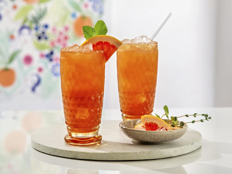Deux verres à long drink Boston jaunes remplis de jus de fruit, garnis de tranches de pamplemousse et de menthe, servis sur un plateau avec un petit bol d’agrumes.