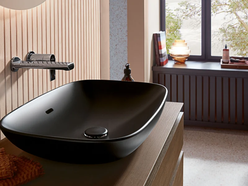 Une salle de bains moderne avec une vasque à poser noire Loop & Friends de Villeroy & Boch, un robinet mural, des touches de bois, une fenêtre avec vue sur l’extérieur et différents articles de toilette.