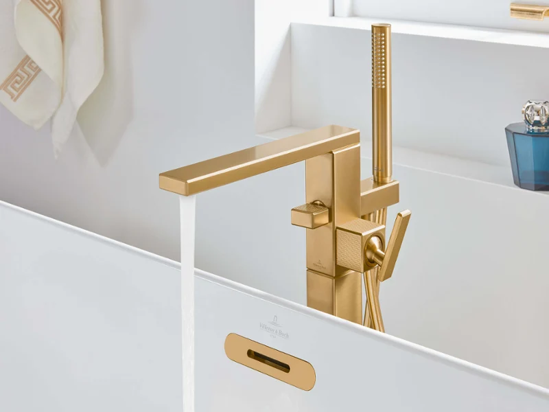 Une robinetterie de baignoire dorée Mettlach avec douchette à main Une salle de bains moderne avec un robinet doré avec de l’eau qui coule, une serviette accrochée au mur et des objets décoratifs sur l’étagère.