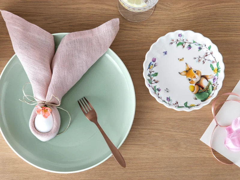 Une assiette verte "it’s My Match" de Villeroy & Boch et une assiette florale décorative Spring Fantasy sur une table en bois. Une assiette verte "it’s My Match" de Villeroy & Boch, décorée avec une serviette rose, un œuf blanc, une fourchette marron et une assiette florale décorative Spring Fantasy avec une figurine de lapin sur une surface en bois.