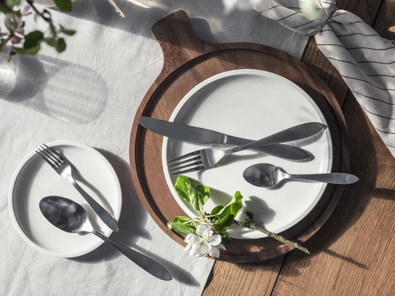 Une table dressée avec des assiettes blanches sur lesquelles sont disposés des couverts Arthur. Une table dressée avec deux assiettes blanches, des couverts Arthur disposés dessus et une branche de fleur sur une nappe ensoleillée.