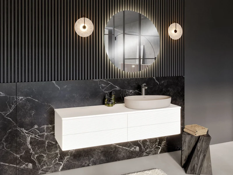 Une salle de bains moderne avec un plan de toilette suspendu, un miroir rond, deux lampes suspendues et des murs structurés sombres. Un lavabo blanc de la collection Antao de Villeroy & Boch et une décoration minimaliste sont présents, ainsi qu’une petite table et un tapis.