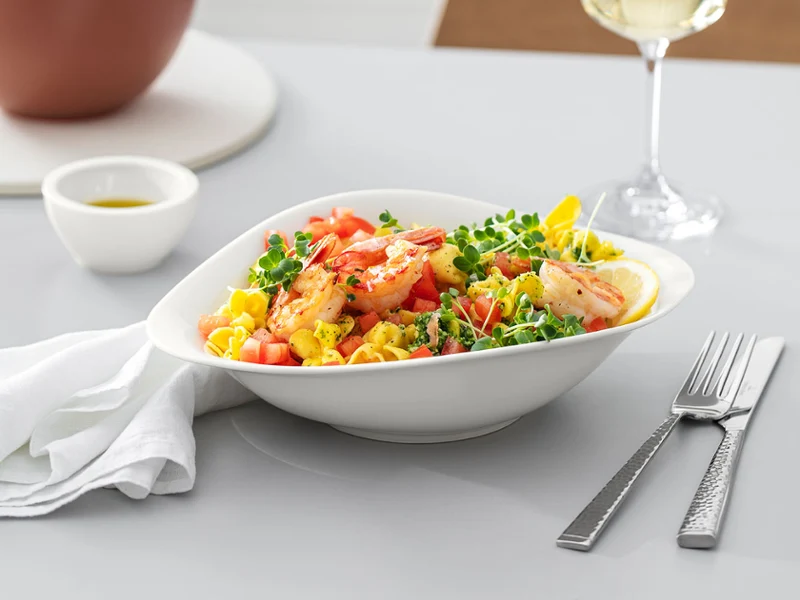 Une assiette Vapiano de Villeroy & Boch remplie de pâtes et de salades sur une table en bois Une assiette Vapiano de Villeroy & Boch remplie de salade de pâtes sur une table en bois Sur la table, un verre à vin et des couverts de table Villeroy & Boch