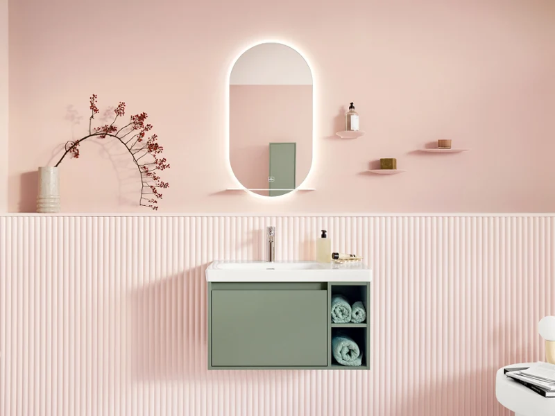 Espace de lavage Skyla tendance et aménagé avec style dans des tons pastel. L'espace de lavage de cette salle de bain aménagée avec style est présenté en vue frontale et se compose d'un meuble-lavabo avec meuble bas assorti et robinetterie chromée - tous deux de la collection de salle de bain Skyla - ainsi que d'un miroir ovale de la collection More To See Lite+.