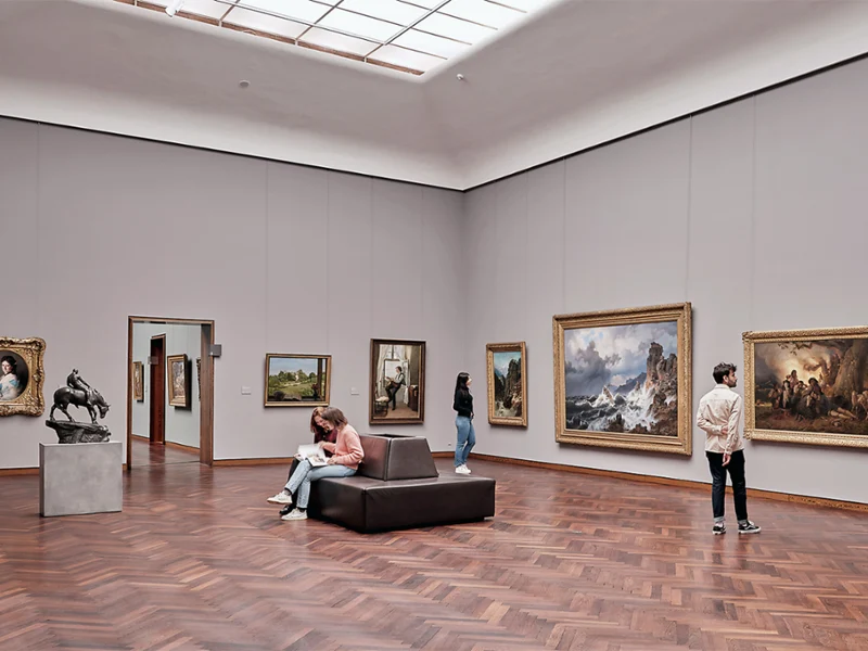 Quatre personnes regardent des tableaux et une sculpture dans une galerie d'art spacieuse avec des parquets et de hauts plafonds.