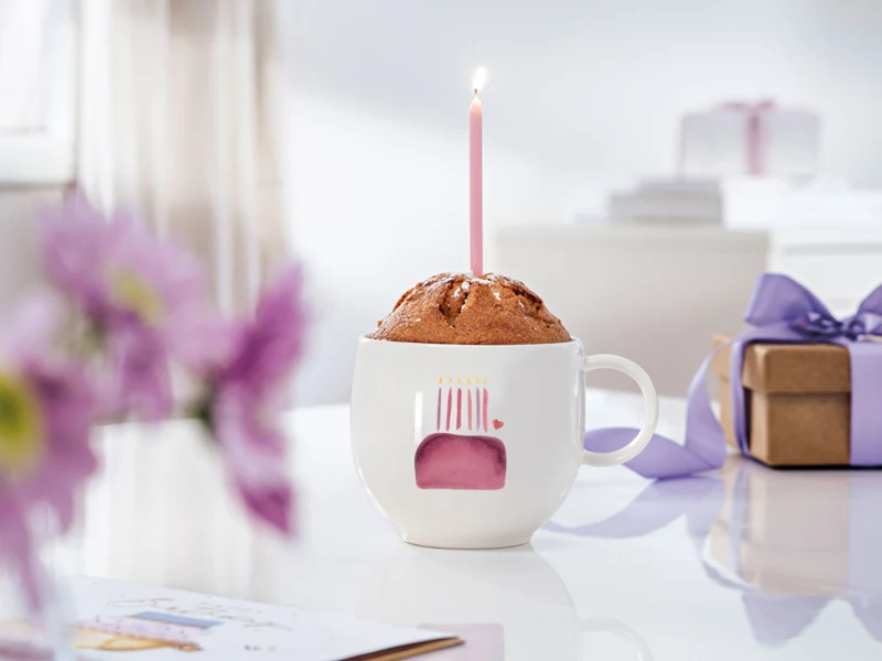Un mugcake avec des bougies dans une tasse With Love Birthday sur une table Un mugcake avec des bougies dans une tasse With Love Birthday sur une table avec des fleurs violettes et une tasse dans une boîte cadeau beige.