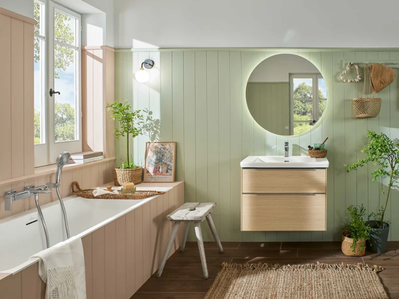 Une salle de bains avec une baignoire Subway 3.0 de Villeroy & Boch, un miroir rond et un plan de toilette en bois. Les plantes et les décorations en osier apportent une touche naturelle. Des boiseries aux couleurs pastel soulignent l’esthétique rustique. La lumière entre par la fenêtre.