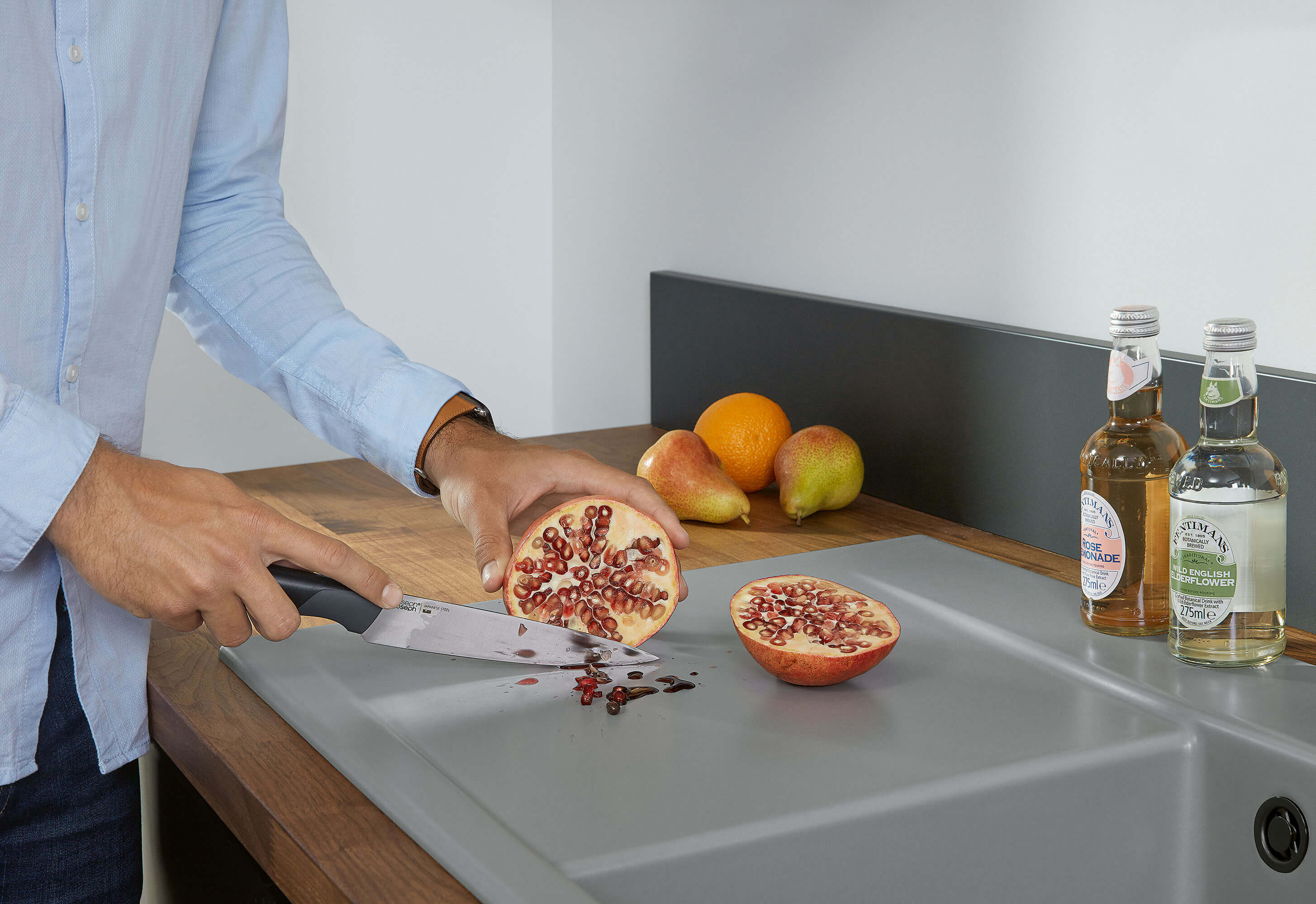 Sur un comptoir de cuisine, une personne découpe une grenade avec un couteau. En arrière-plan, on voit des bouteilles et différents fruits.