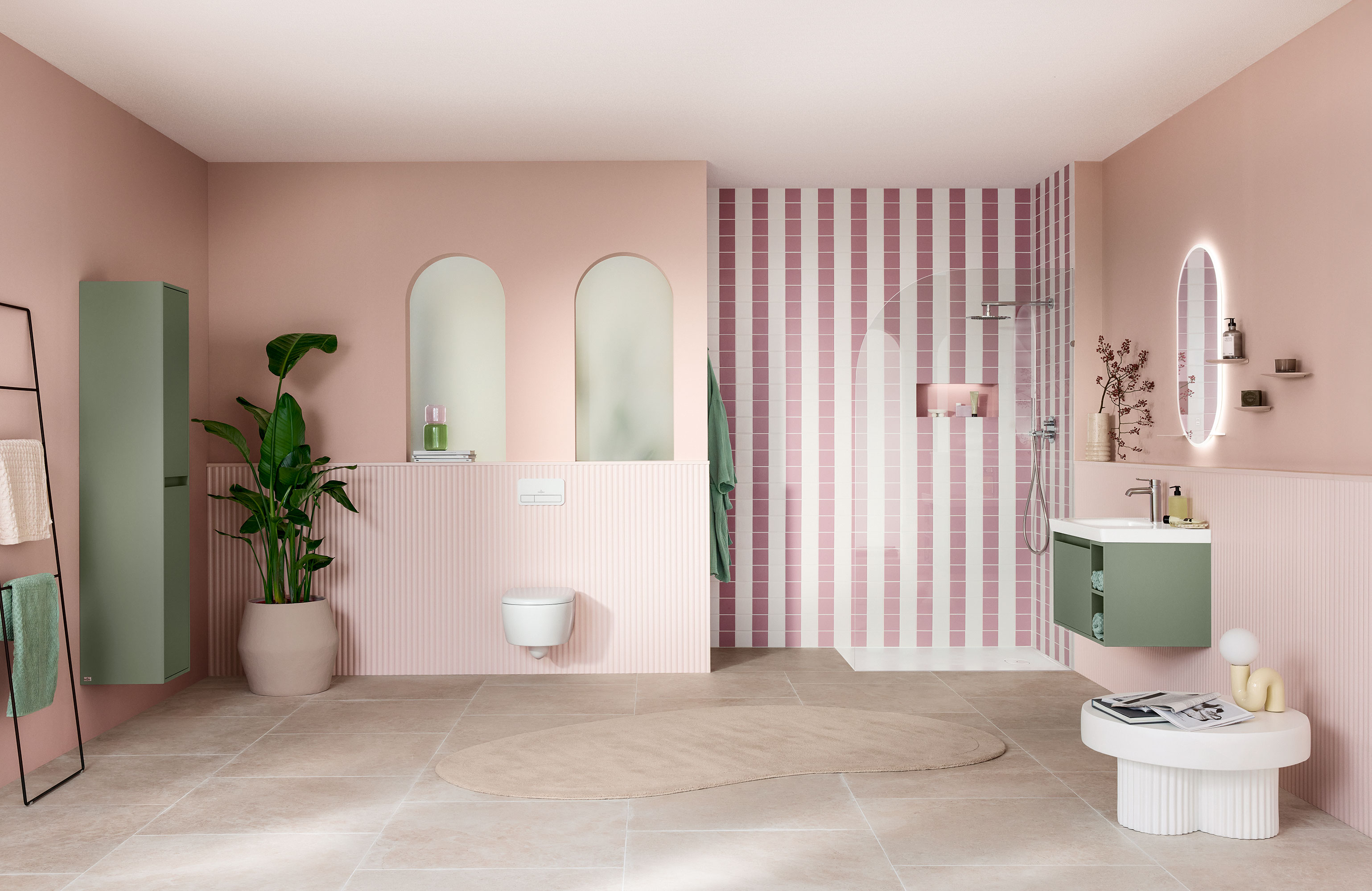 Meuble sous-vasque vert Skyla dans une salle de bains rose pastel Salle de bains moderne avec des murs roses, un espace douche rayé, des touches de vert, de grands miroirs et une plante en pot.