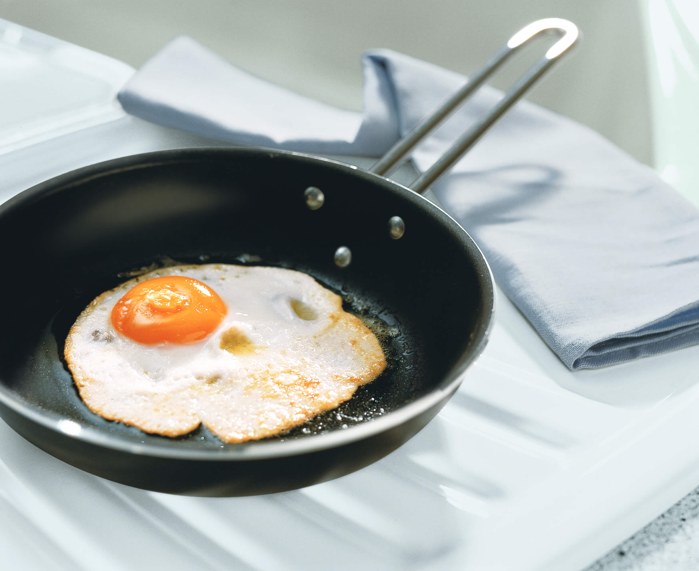 Un œuf au plat est cuit dans une poêle antiadhésive, à côté de laquelle se trouve un torchon plié.