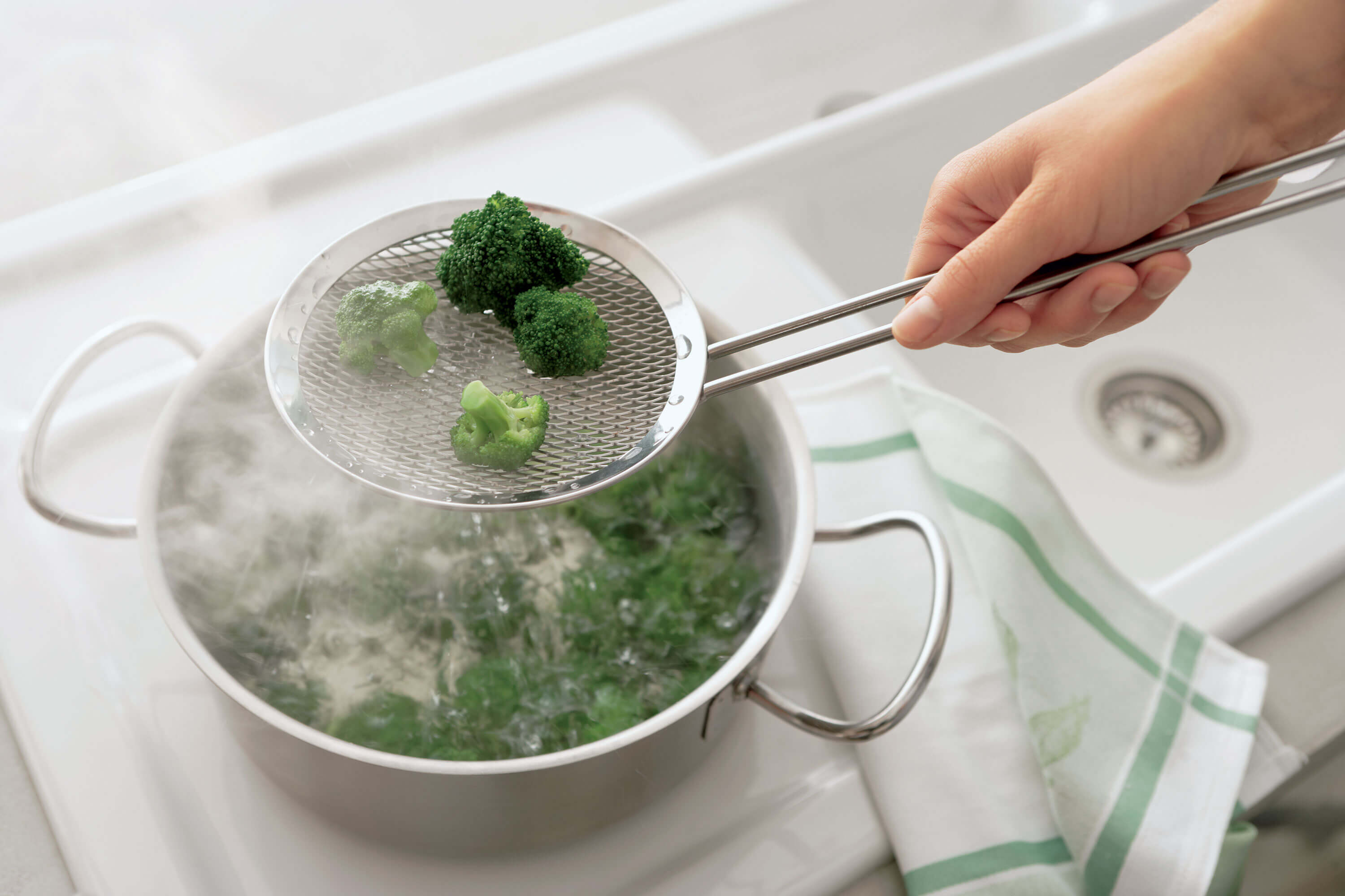 Une personne tient une écumoire remplie de bouquets de brocoli au-dessus d’une casserole d’eau bouillante sur le feu, dans laquelle se trouvent d’autres brocolis. Un essuie-main rayé est posé sur le plan de travail près de l’évier.