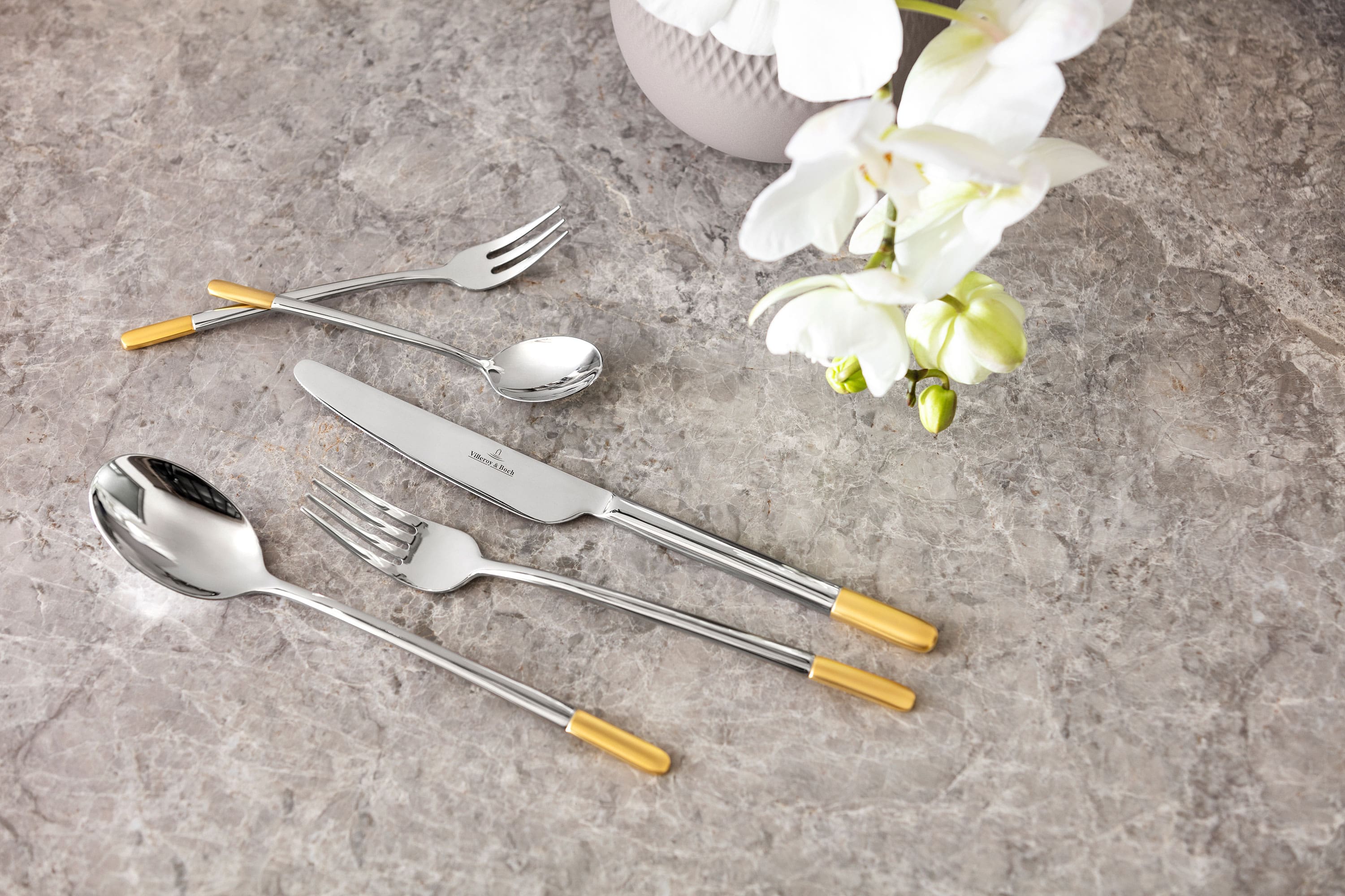 Un ensemble de couverts de table Ella de Villeroy & Boch avec des notes dorées Un élégant ensemble de couverts de table Ella avec des touches dorées sur un plateau de table en marbre, accompagné d’une orchidée blanche.