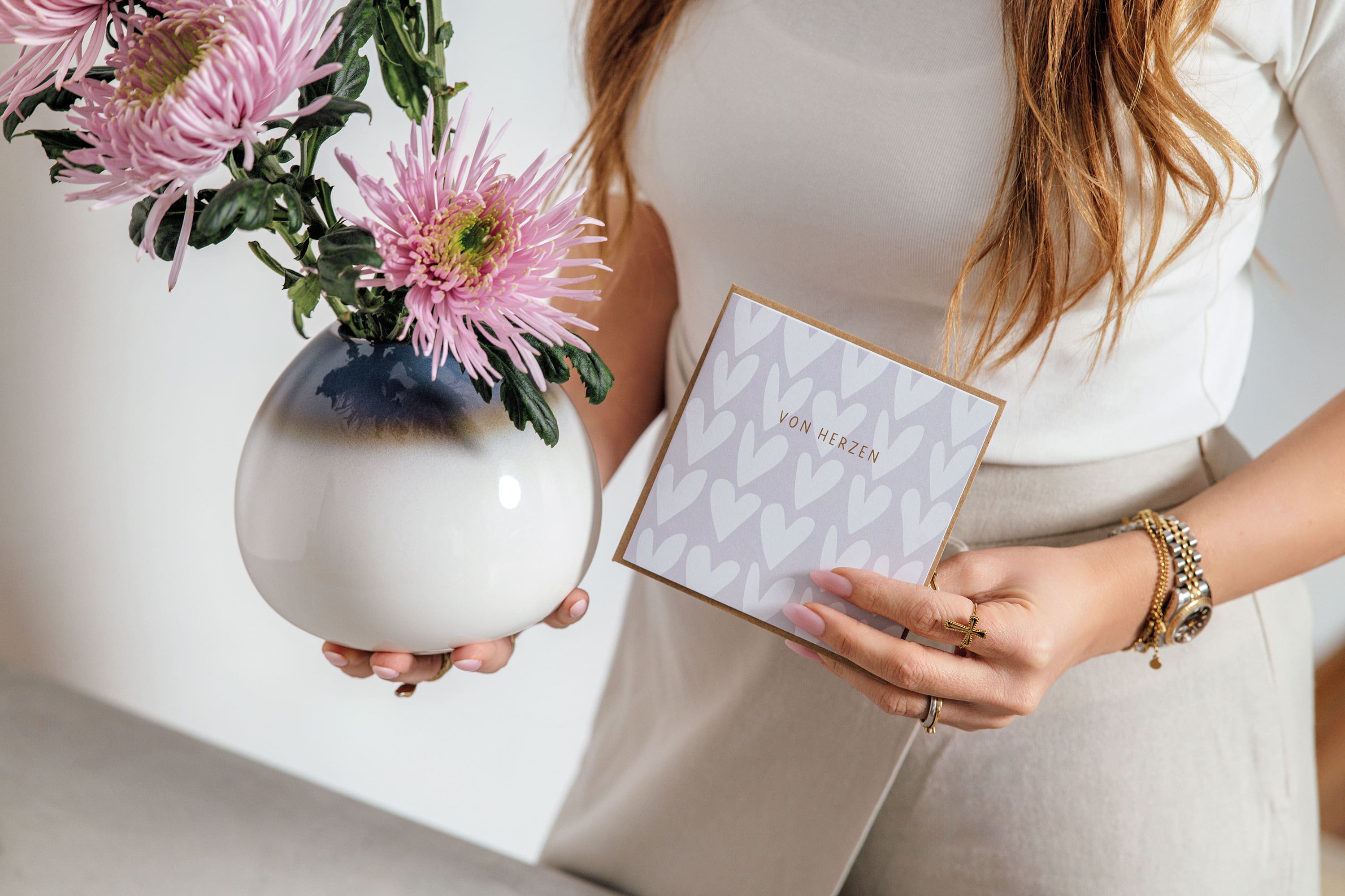 Une femme tient une carte portant l’inscription « von Herzen » et un vase Lave avec des fleurs roses et se tient à côté d’une table.